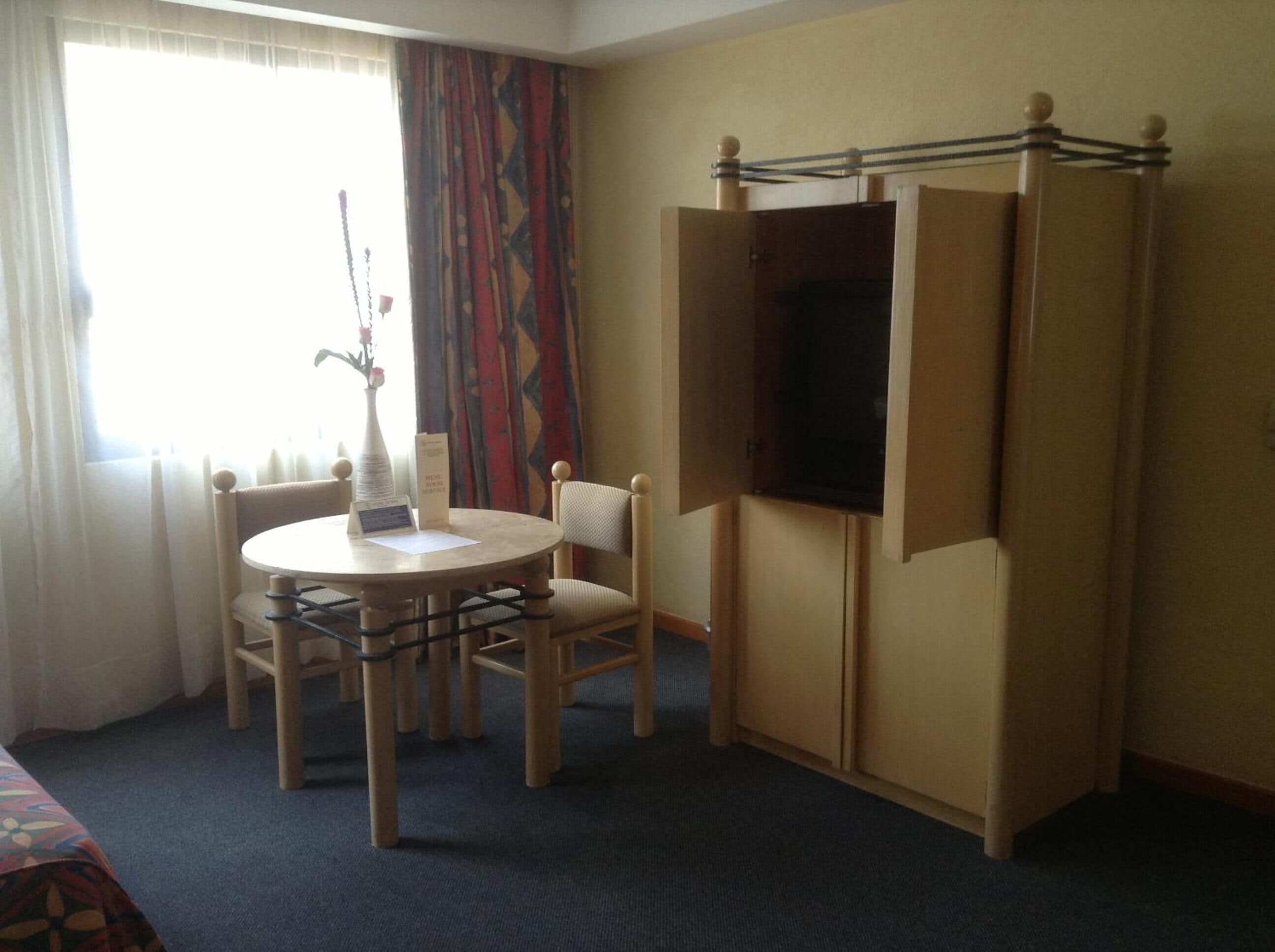 Quarto de hotel com uma mesa redonda, duas cadeiras, uma televisão e um guarda-roupa.