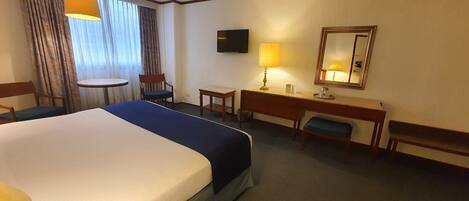 Habitación de hotel con una cama grande, un escritorio, una silla, un televisor, una lámpara y un espejo.