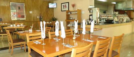 Um restaurante com mesas e cadeiras de madeira, um balcão com uma máquina de café e uma televisão na parede.