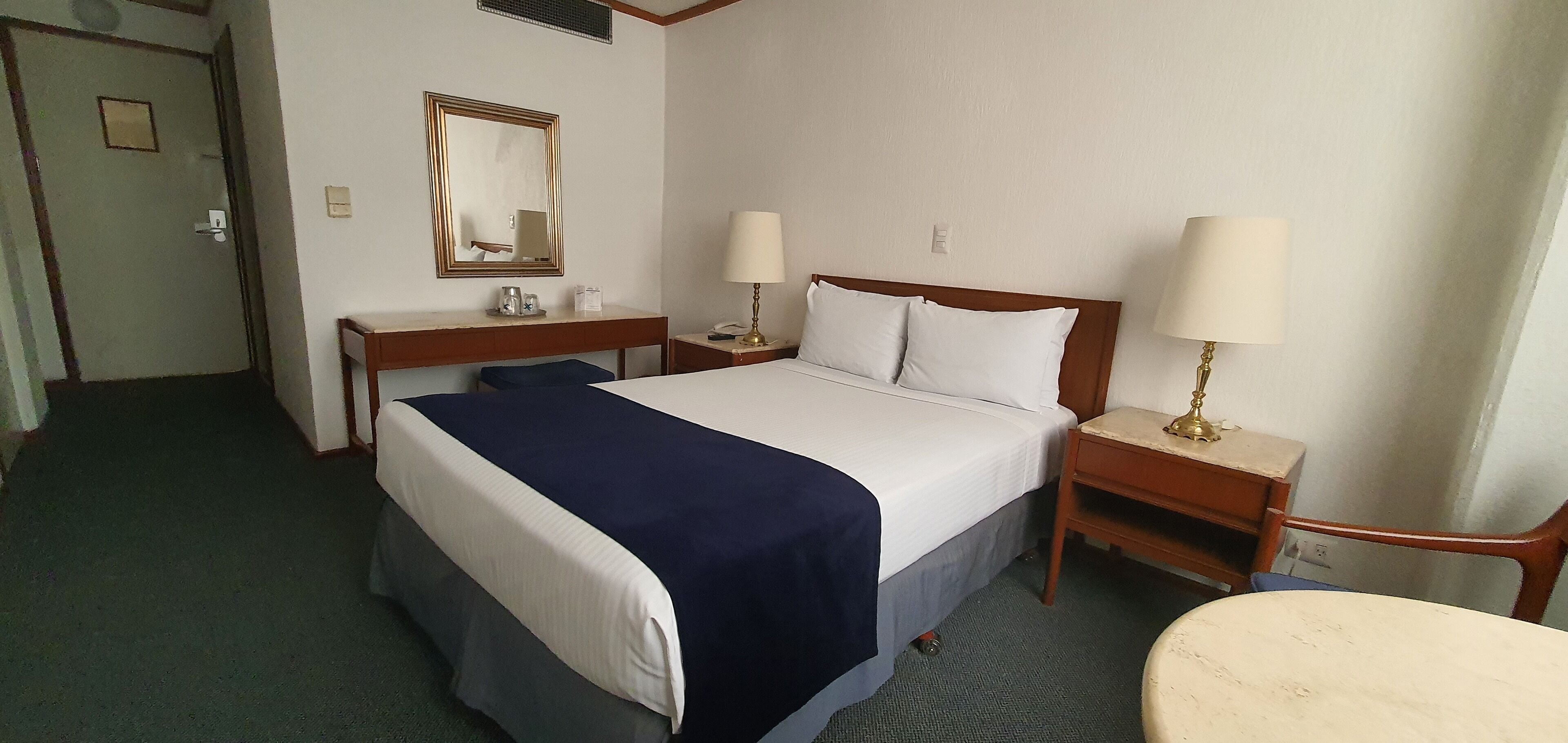 A hotel room with a large bed, two bedside tables with lamps, a desk with a chair, a mirror, and a door.