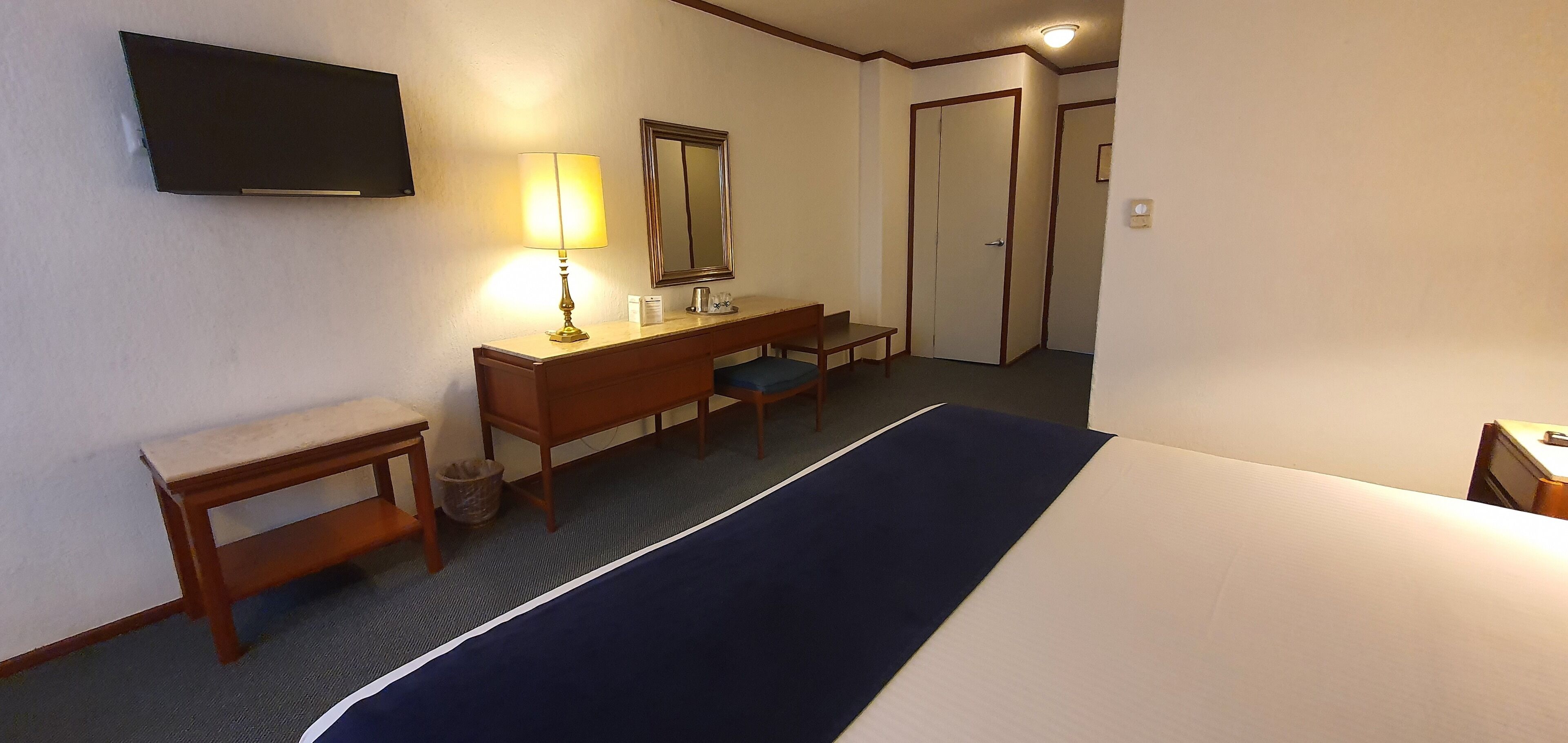 Una habitación de hotel con una cama, un escritorio, una silla, una lámpara, un espejo y un televisor en la pared.
