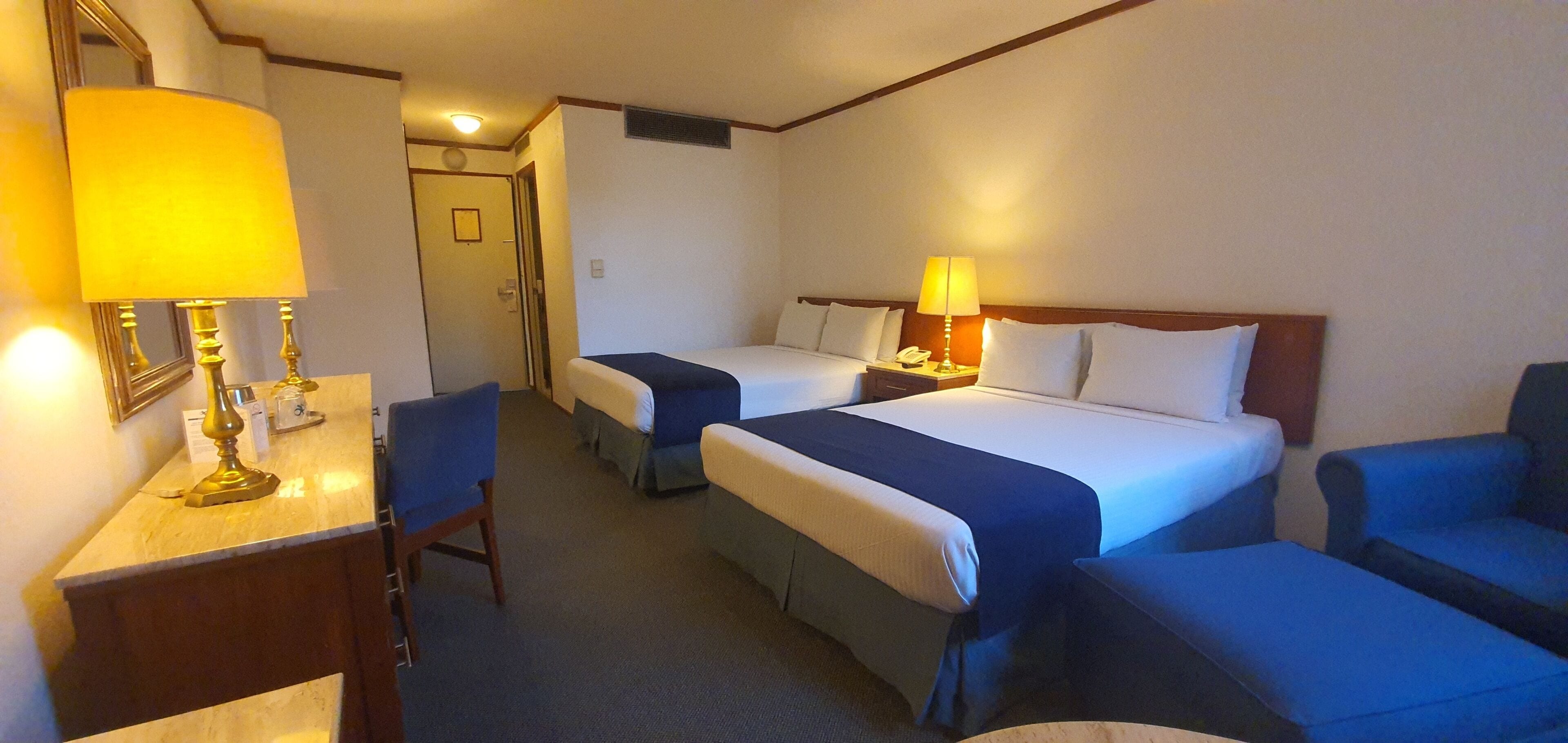 A hotel room with two beds, a desk with a lamp, and a chair.