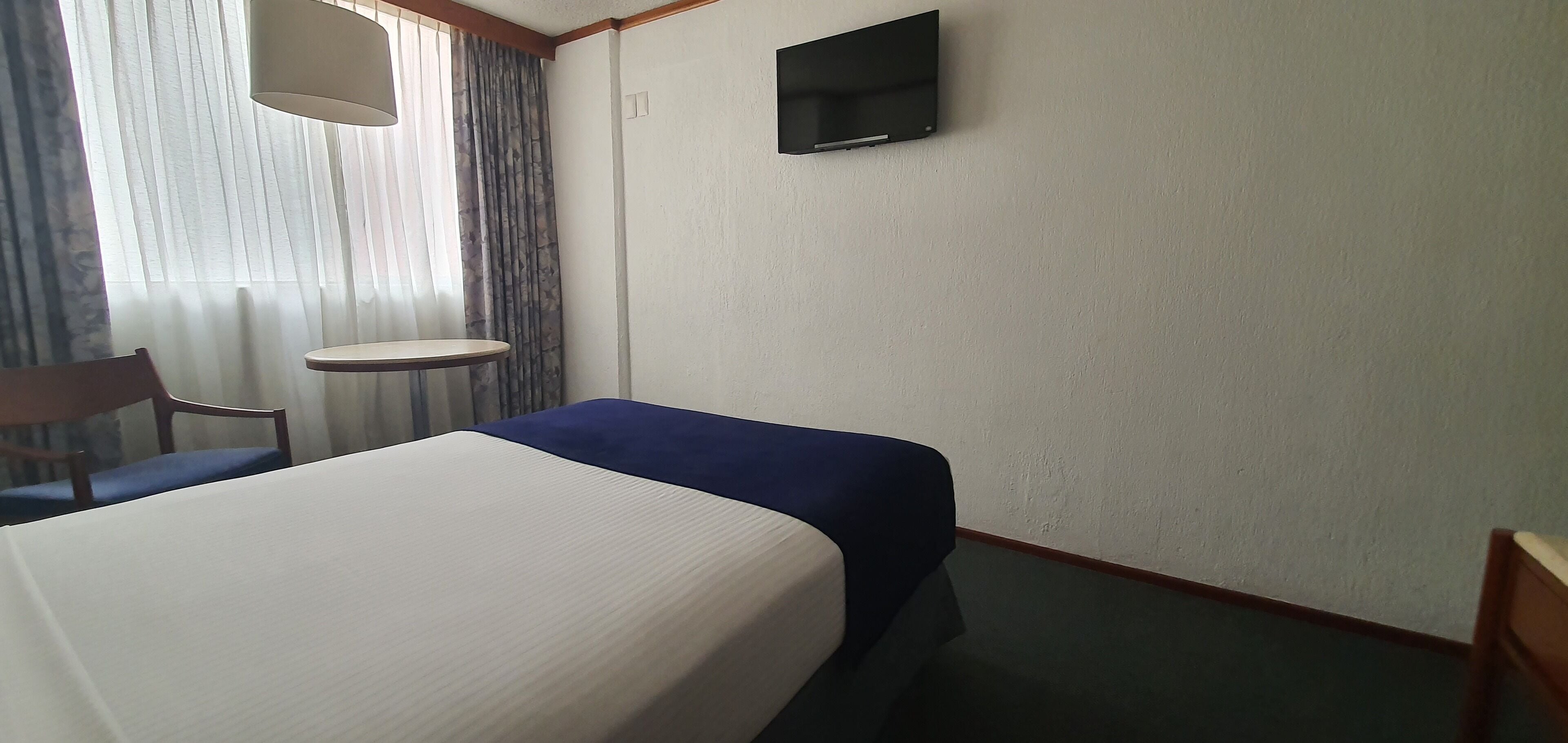 Een hotelkamer met een bed, een klein tafeltje, een stoel, een aan de muur bevestigde televisie en een raam met gordijnen.