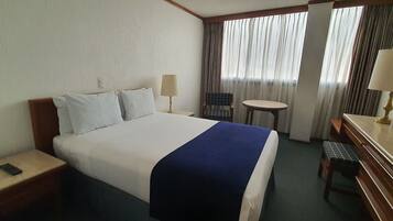 A hotel room with a bed, a desk, a chair, a small table, and a lamp.