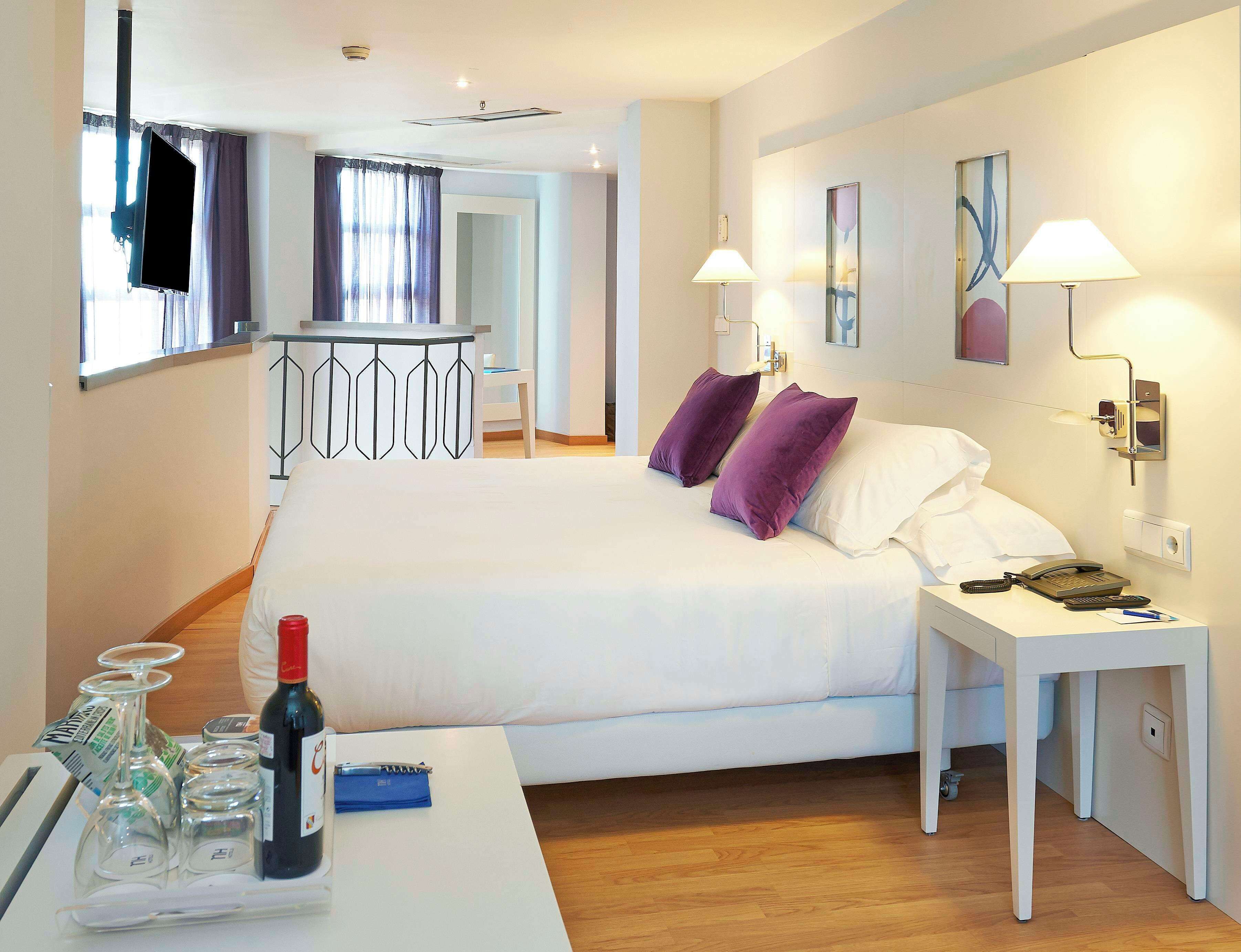 A hotel room with a bed, bedside table, lamp, and a small desk with a telephone and a bottle.