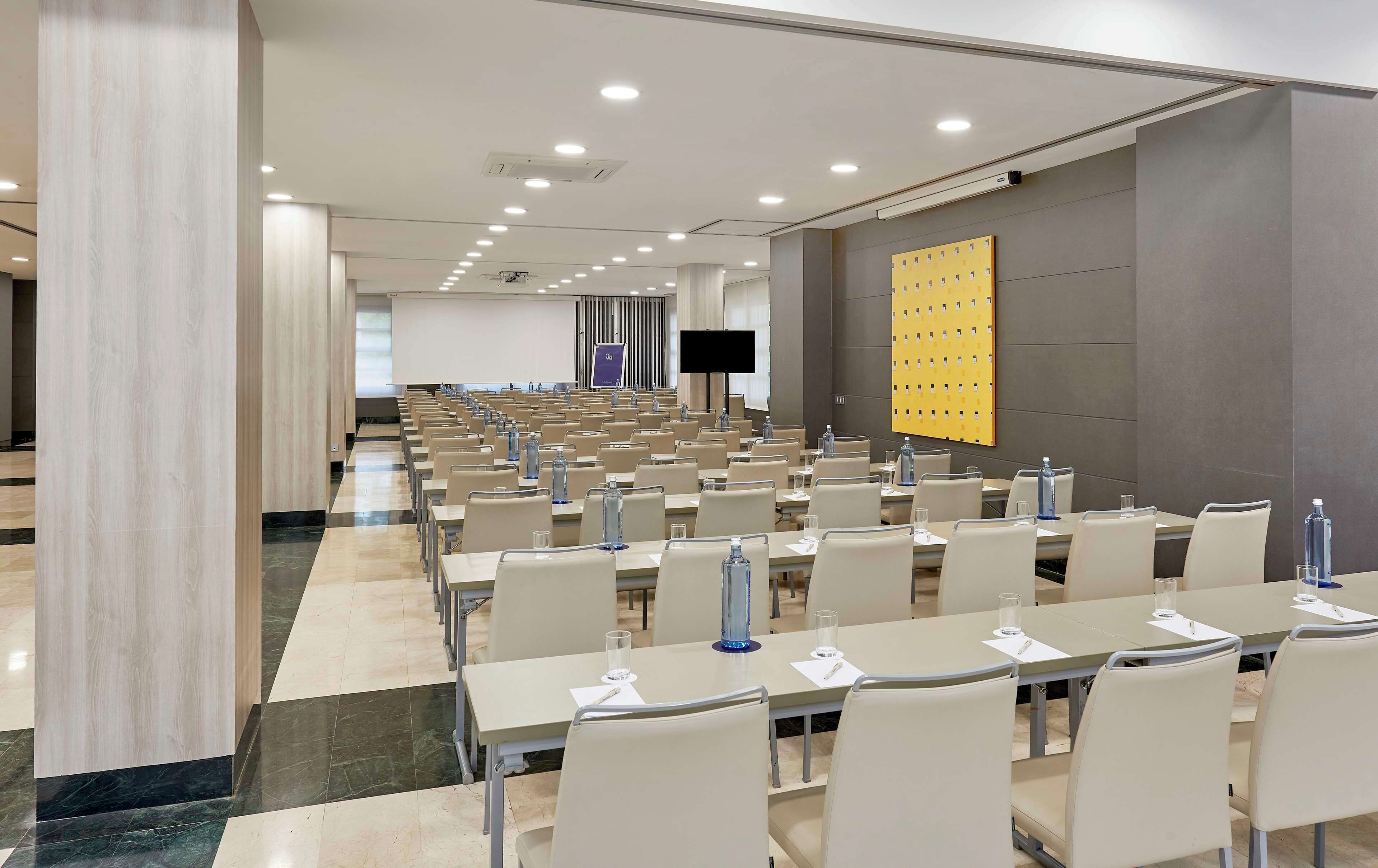 Une salle de conférence avec des rangées de tables et de chaises, un grand écran et un projecteur.