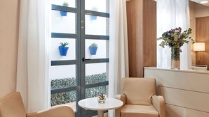 A hotel room with two beige armchairs, a small round table, a vase of flowers, and a window with a view of a balcony with potted plants. - NH Valencia Las Ciencias Hotel (Valencia)