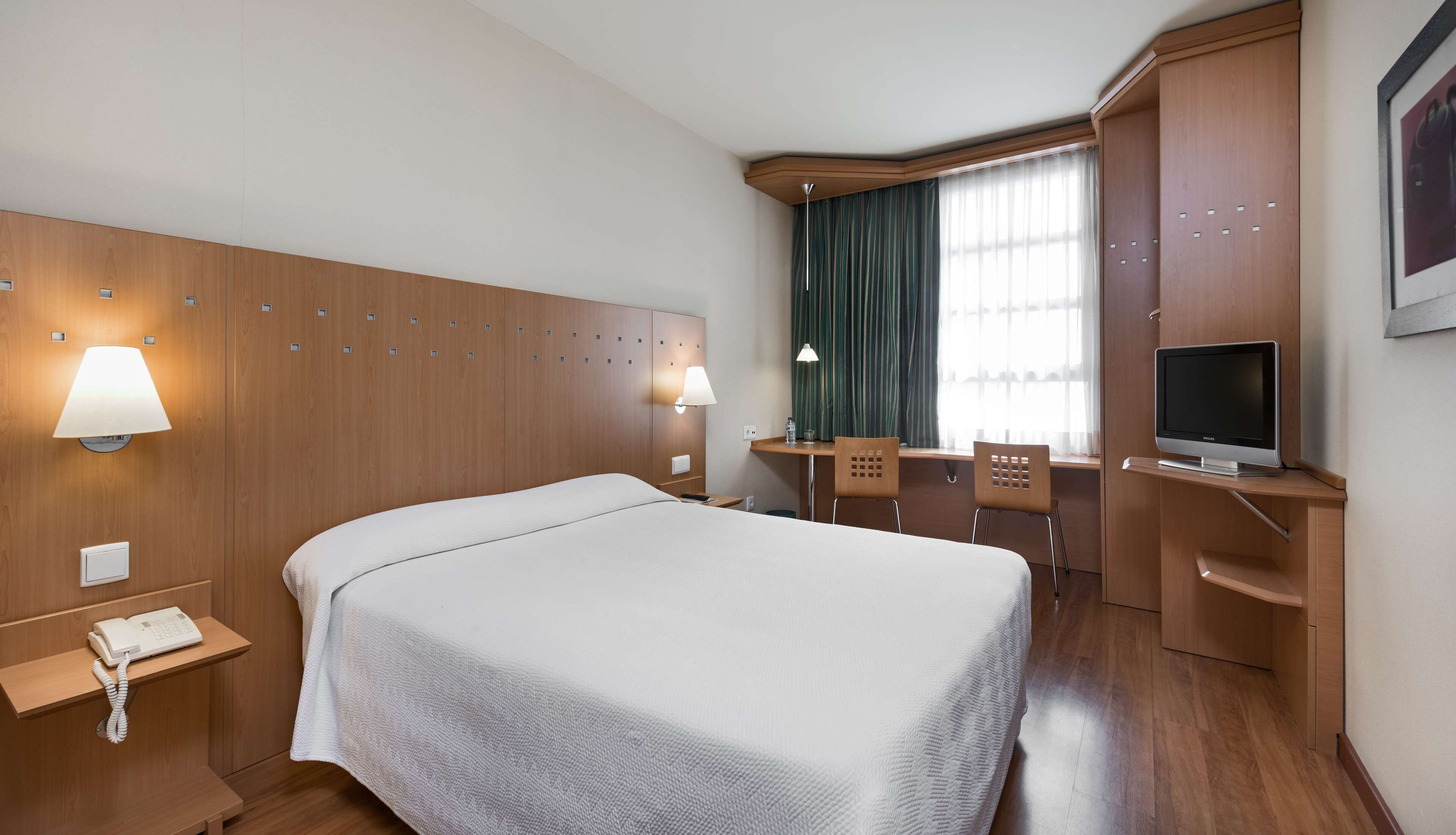 Una habitación de hotel con una cama grande, un escritorio con silla, televisor y ventana con cortinas.