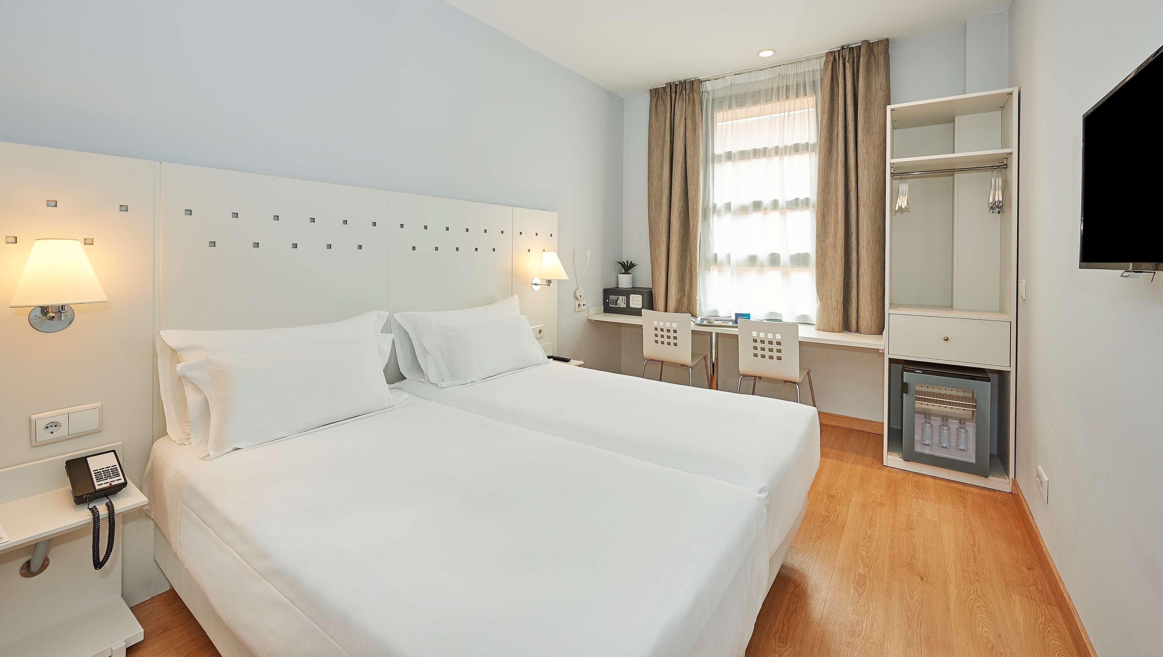 Une chambre d’hôtel avec un grand lit, un bureau avec une chaise, un petit réfrigérateur, un miroir et une télévision.