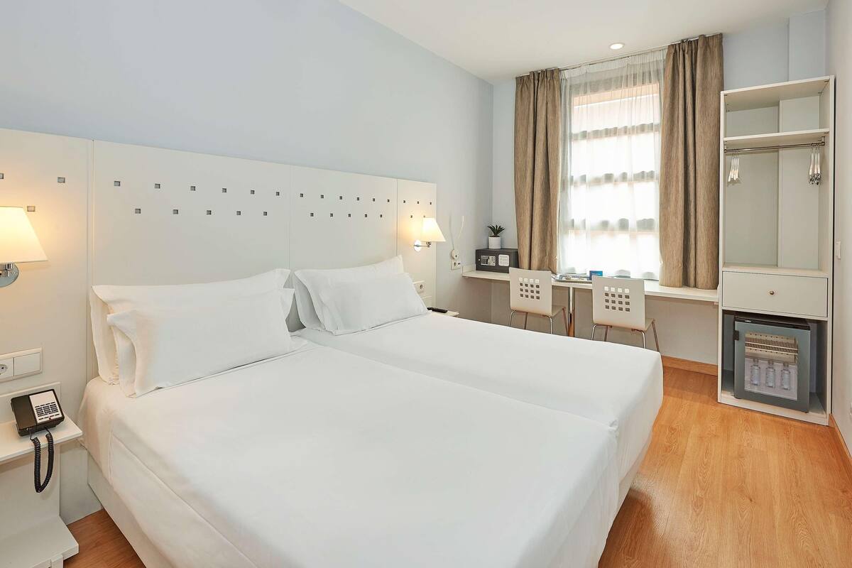 Une chambre d’hôtel avec un grand lit, un bureau avec une chaise, un petit réfrigérateur, un miroir et une télévision.