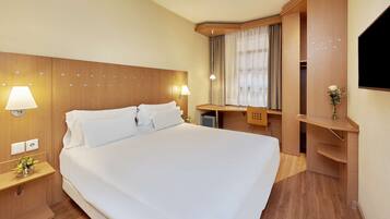 Habitación de hotel con una cama grande, lámparas de noche, un escritorio con silla, un ventanal con cortinas y un televisor montado en la pared.