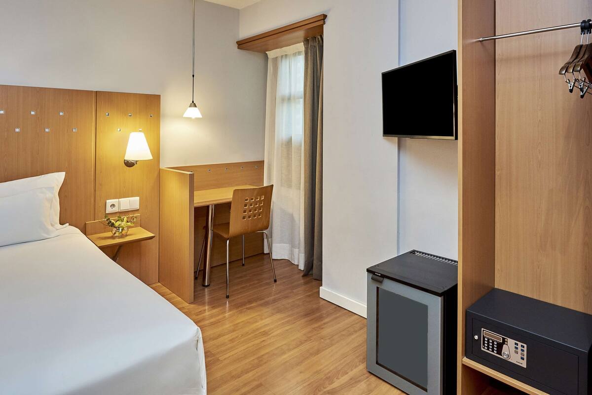 A hotel room with a bed, desk, chair, TV, and a small heater.