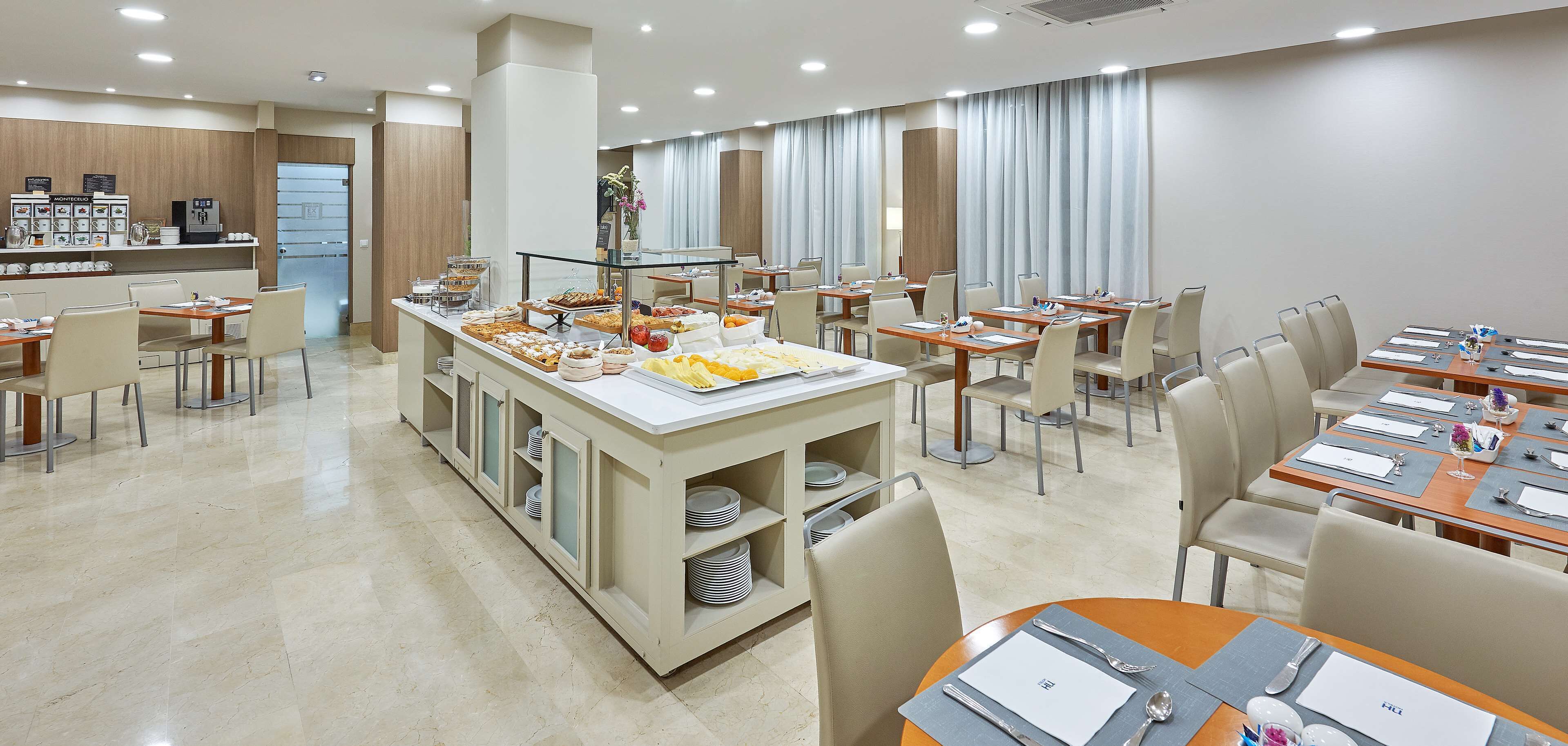 Un comedor de hotel con estaciones de buffet, mesas dispuestas para comer y un amplio ventanal con cortinas.
