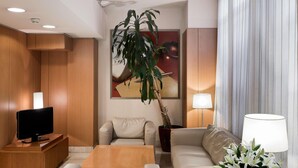 A modern living room with a sofa, a coffee table, a TV on a wooden stand, a potted plant, and a lamp on a side table. - NH Valencia Las Ciencias Hotel (Valencia)