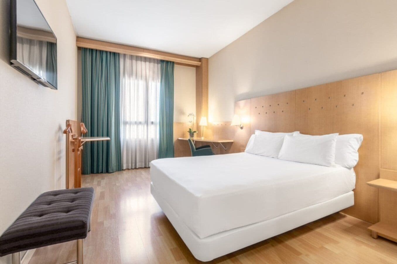 A hotel room with a large bed, a wooden headboard, a desk with a chair, a small table, and a bench.