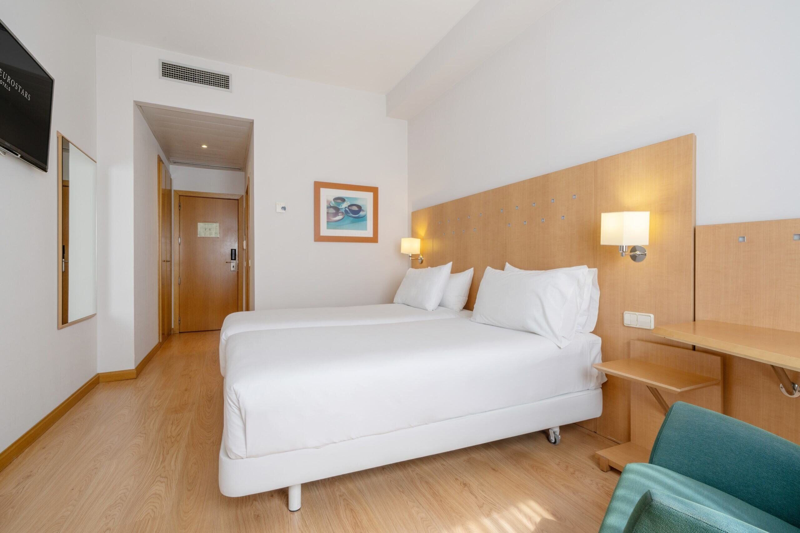 A hotel room with a single bed, bedside table, lamp, and a television on the wall.