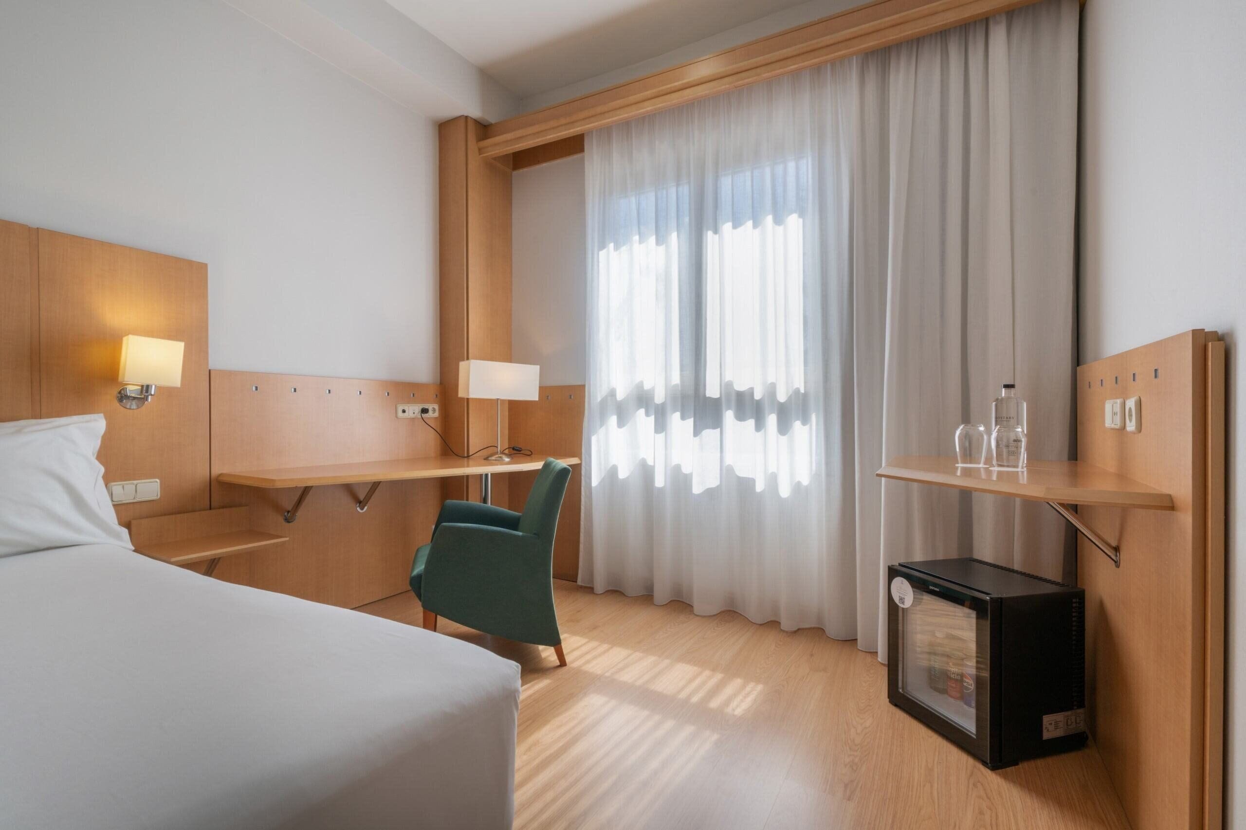 A hotel room with a bed, a desk, a chair, a small refrigerator, and a window with curtains.
