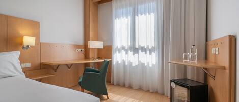 Une chambre d’hôtel équipée d’un lit, d’un bureau, d’une chaise, d’un petit réfrigérateur et d’une fenêtre avec des rideaux.