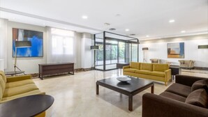 A spacious lobby with a yellow sofa, a brown sofa, and a coffee table. There is a glass partition and a large window with a view of trees. - Crisol Leganés (Leganes)