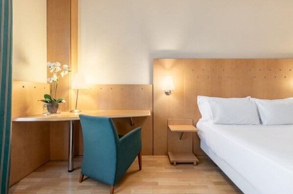 A hotel room with a bed, a desk, a chair, a nightstand, and a wall-mounted lamp. - Crisol Leganés (Leganes)