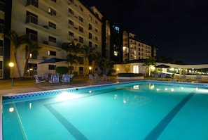 Una piscina con sillas de descanso y sombrillas, rodeada por un edificio de varios pisos con balcones y una entrada bien iluminada.