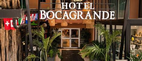 The entrance of Hotel Bocagrade with a modern design, featuring a large sign, potted plants, and a pathway leading to the entrance.