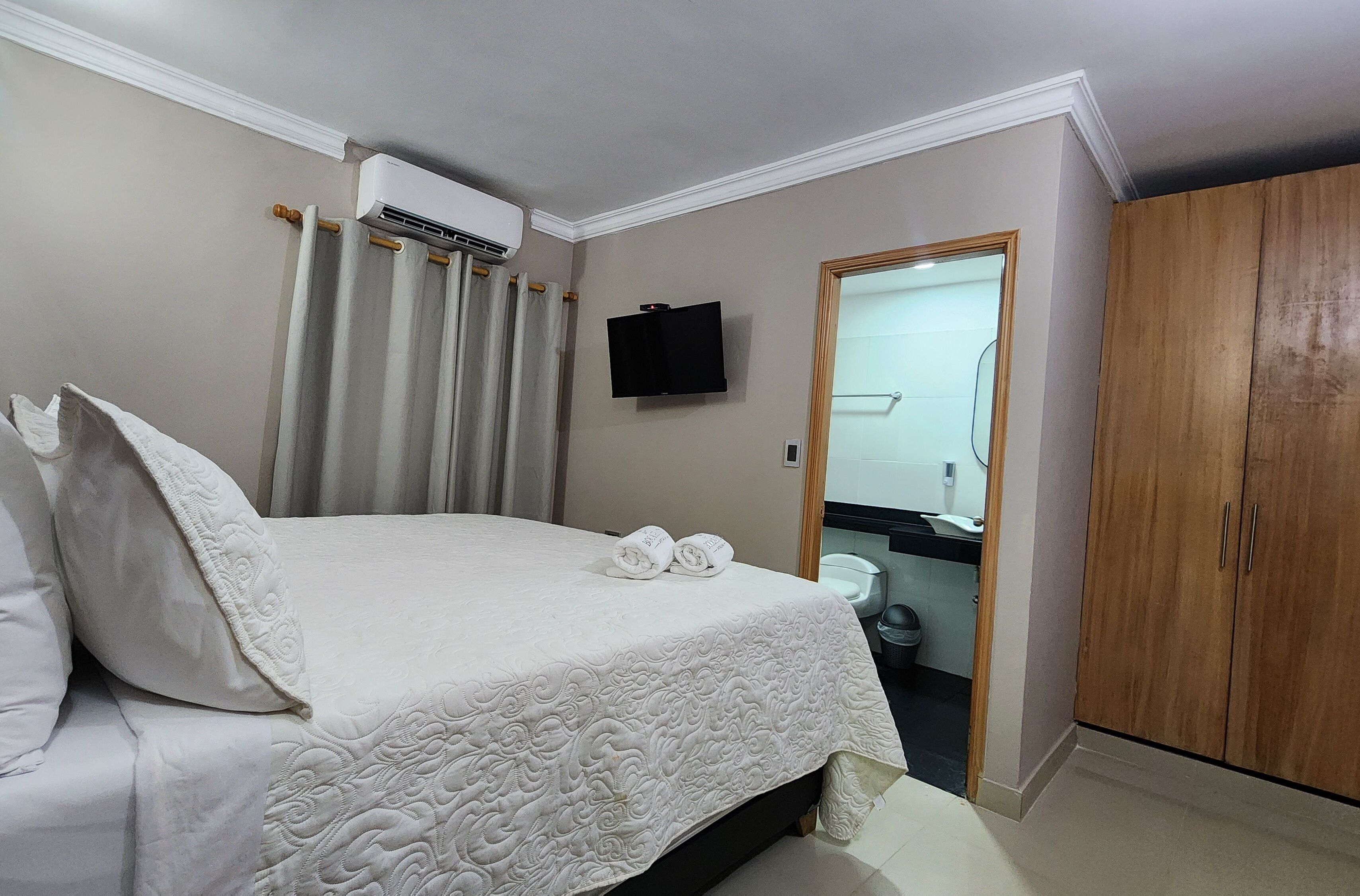 A neatly made bed with a white bedspread, a television mounted on the wall, and a wooden wardrobe.