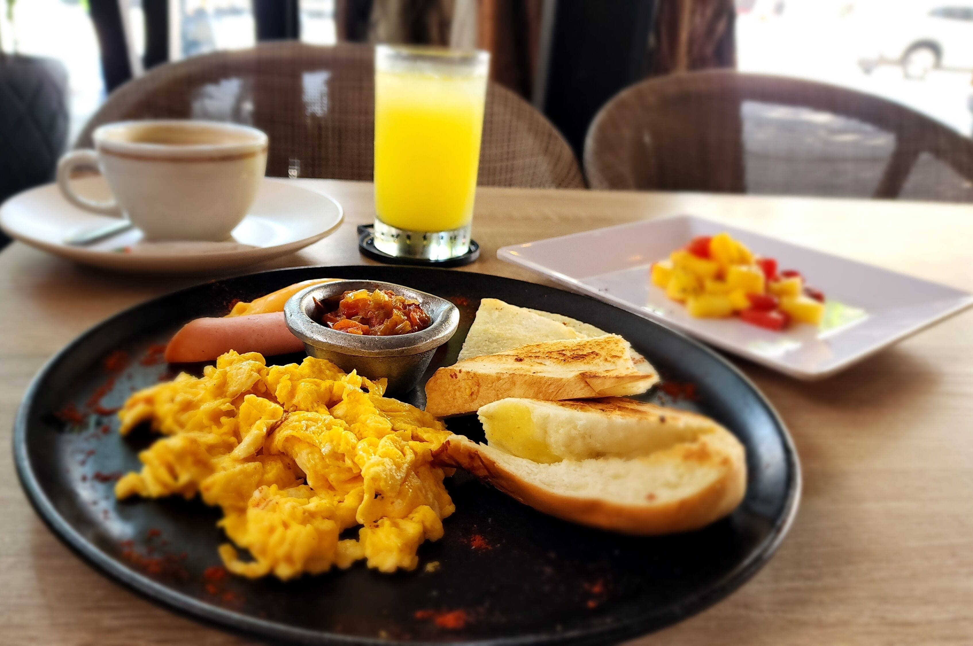 Une assiette de petit-déjeuner avec des œufs brouillés, des toasts et une portion de fruits, une tasse de café et un verre de jus d’orange, le tout posé sur une table.