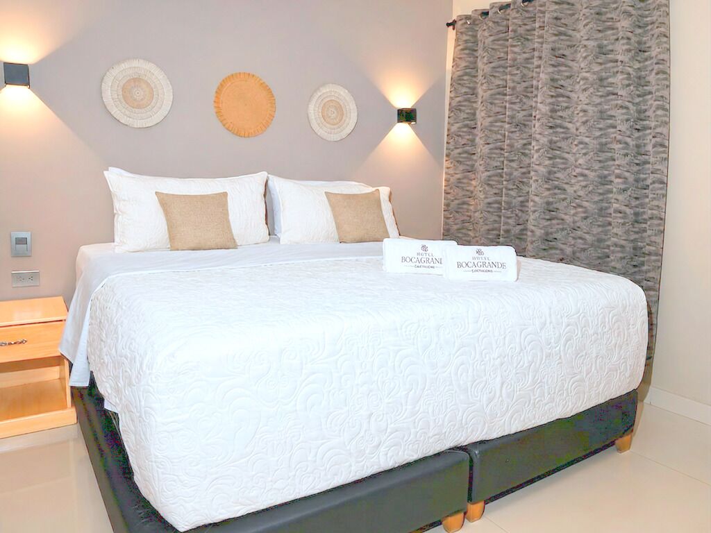 Un lit bien fait, avec une couette blanche et deux oreillers, une table de chevet avec une lampe et une pancarte indiquant le nom de l’hôtel.