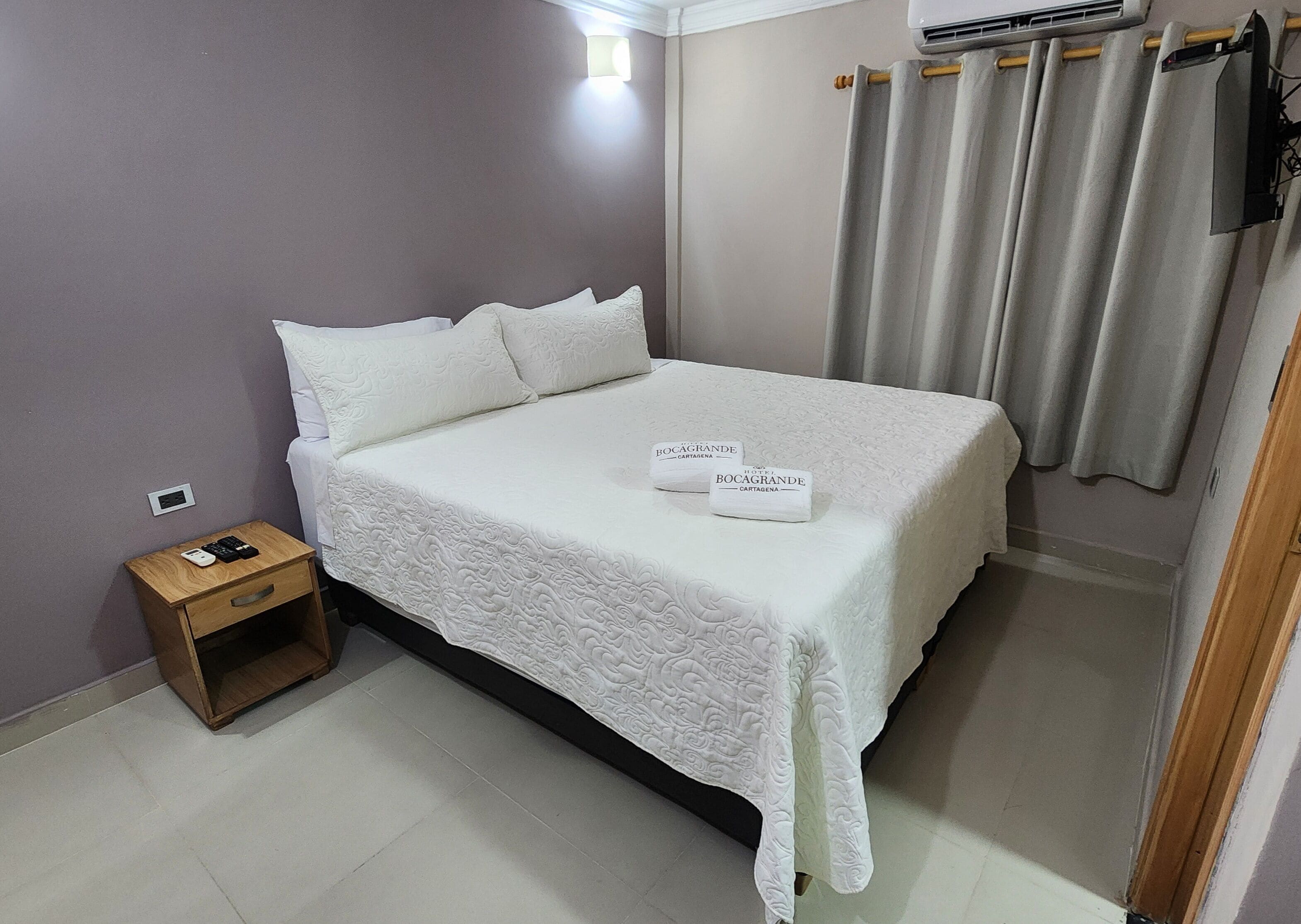 A neatly made bed with a white bedspread and pillows, a small wooden nightstand with a phone and charger, and a wall-mounted air conditioning unit.