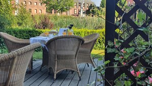 Un espace de restauration en plein air, avec des chaises en osier et une table dressée pour deux, entouré d’une végétation luxuriante et avec un bâtiment en briques en arrière-plan.