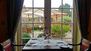 Une table à manger dressée avec des verres à vin et des couverts, une bougie et une plante en pot, donnant sur un jardin et des parasols à l’extérieur.