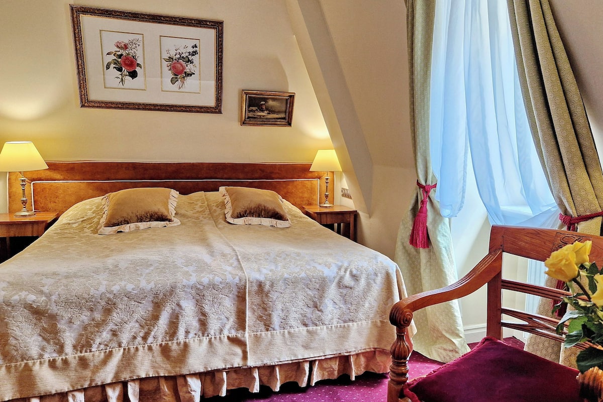 A hotel room with a large bed, two bedside lamps, a wooden headboard, a wooden chair, a vase with yellow flowers, and two framed pictures on the wall.