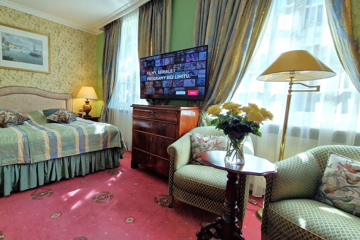 A hotel room with a bed, a television, a desk with a lamp, a chair, and a vase with flowers.