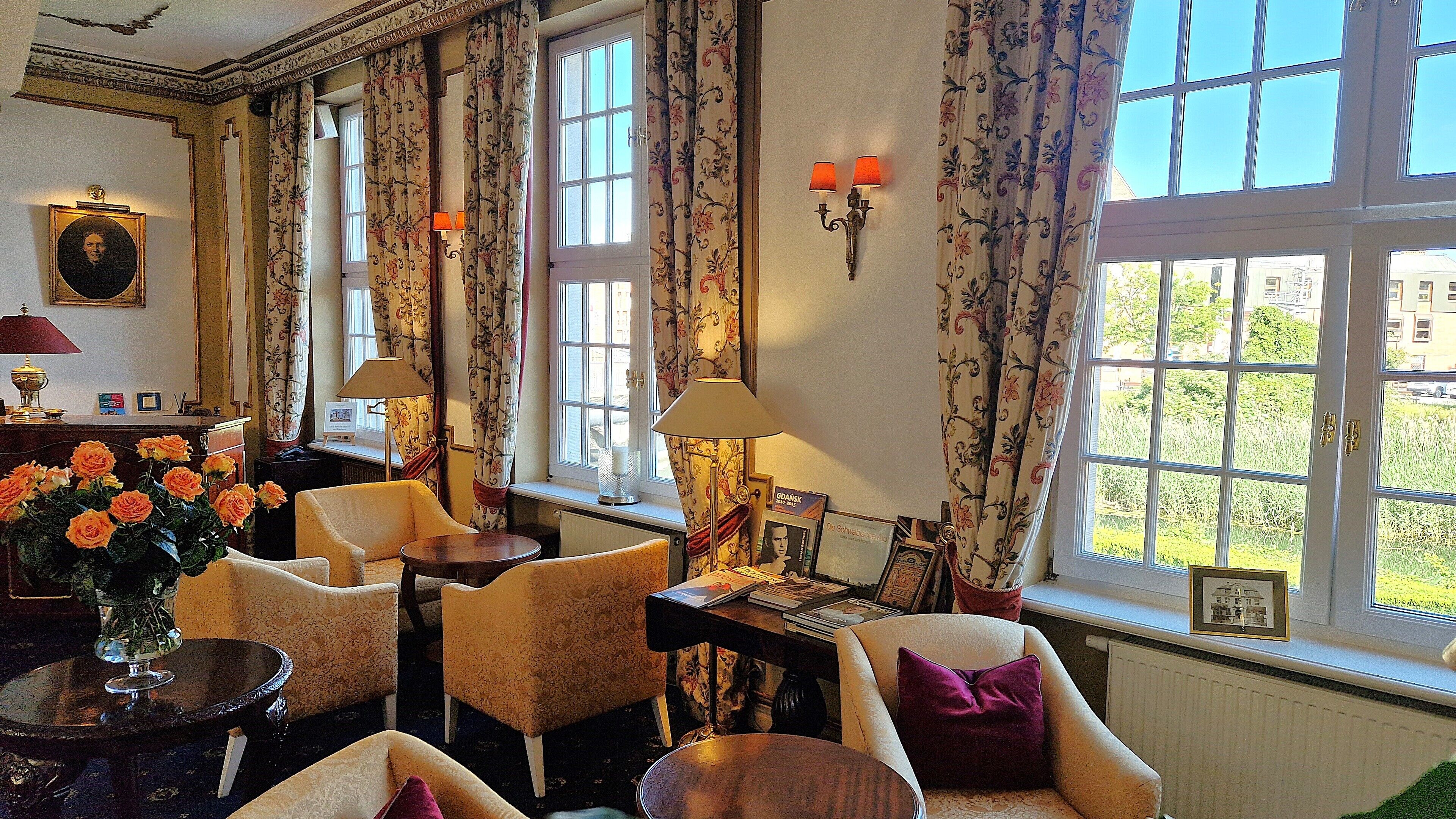 A room with floral curtains, multiple windows, and a view of greenery outside. There are several chairs and a table with a vase of flowers.
