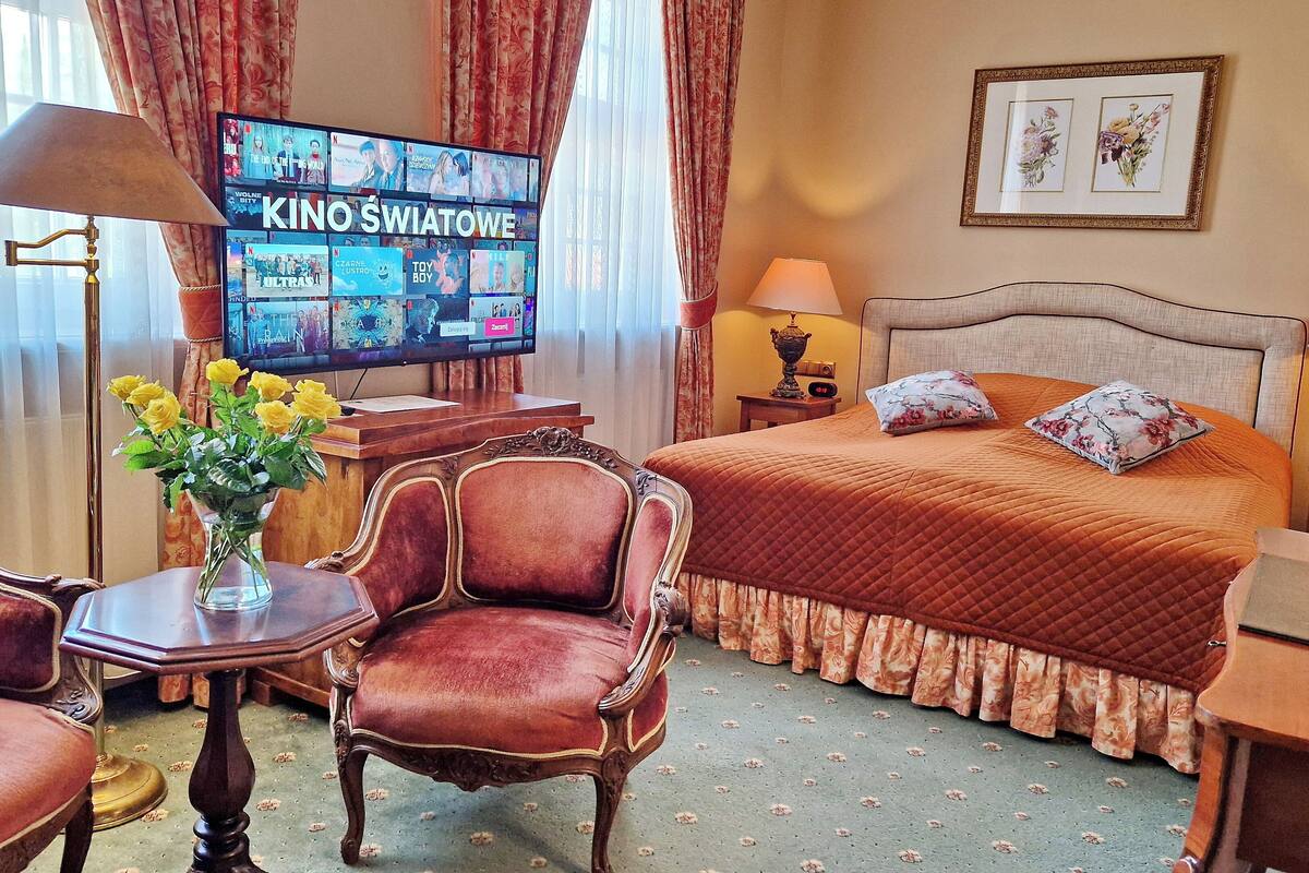 A hotel room with a large bed, two bedside tables with lamps, a desk with a chair, a TV displaying movie channels, and a vase with yellow flowers.