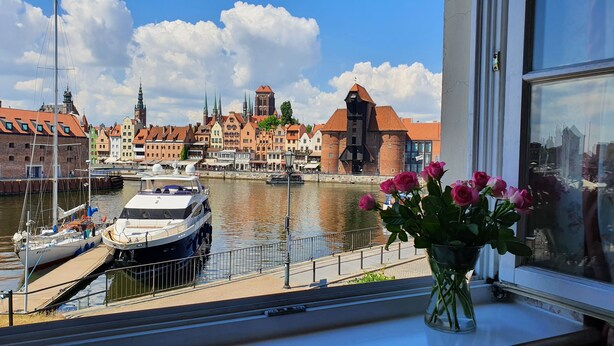 Une vue sur un canal où sont amarrés des bateaux, avec des bâtiments historiques en arrière-plan et un vase de fleurs posé sur un rebord de fenêtre.