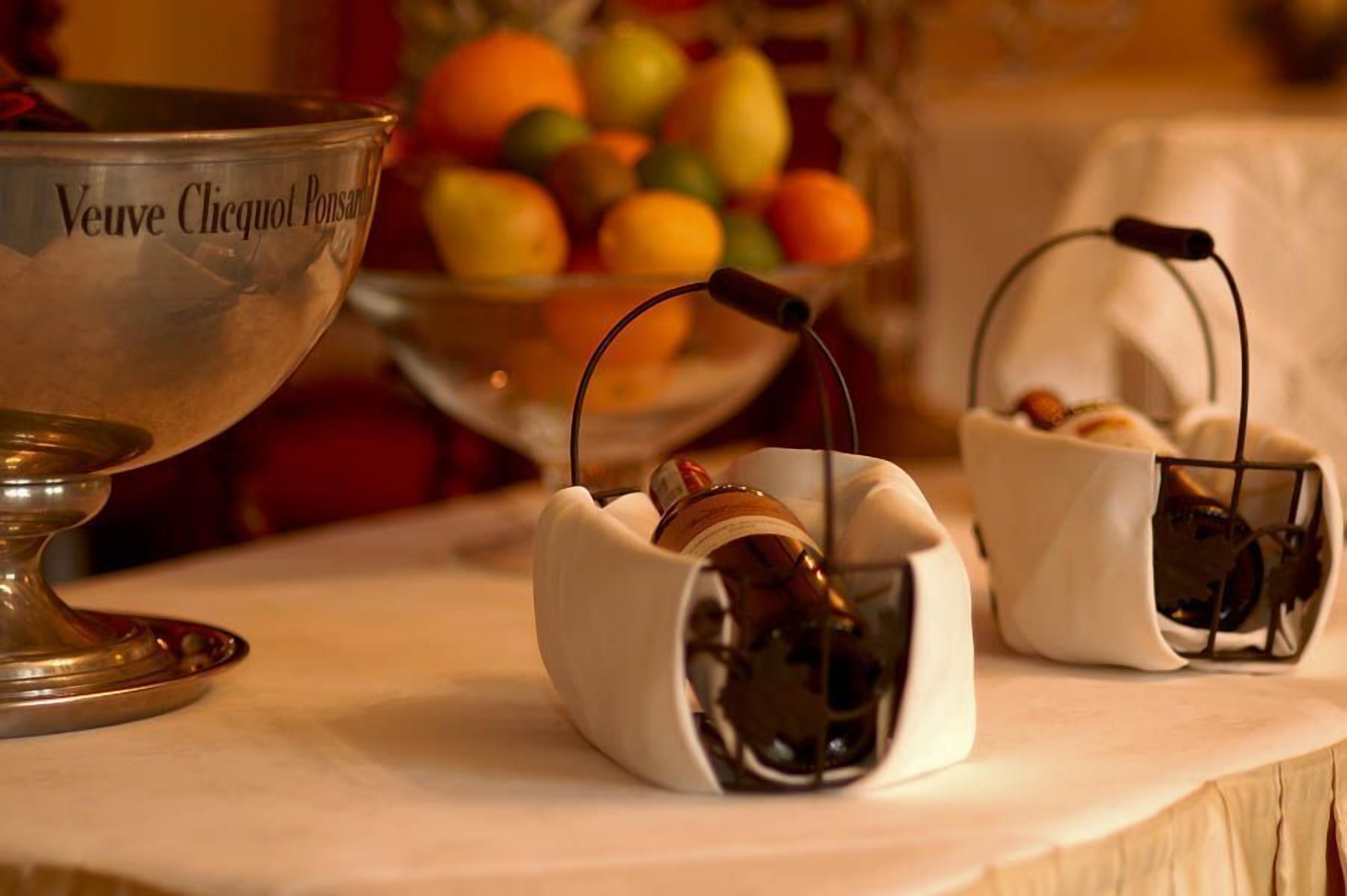 A table with a silver bowl labeled "Veuve Clicquot Ponsard," two small baskets with bottles, and a bowl of fruit in the background.