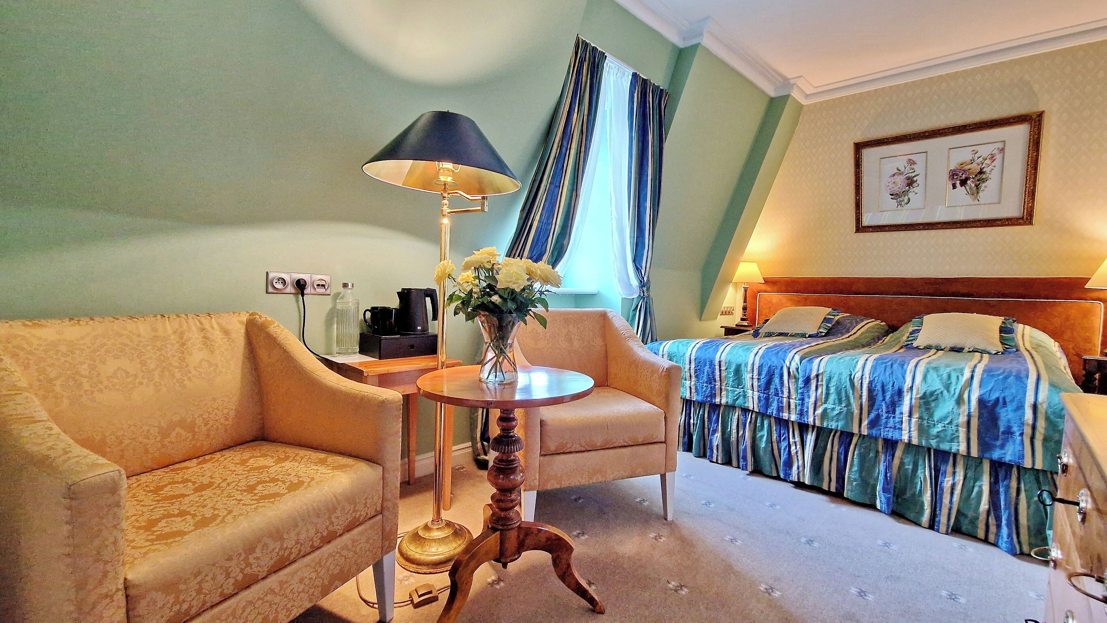 Hotelkamer met een bank, fauteuil, rond tafel met lamp, een bed met gestreept beddegoed en een lijst met bloemistisch kunstwerk aan de muur.