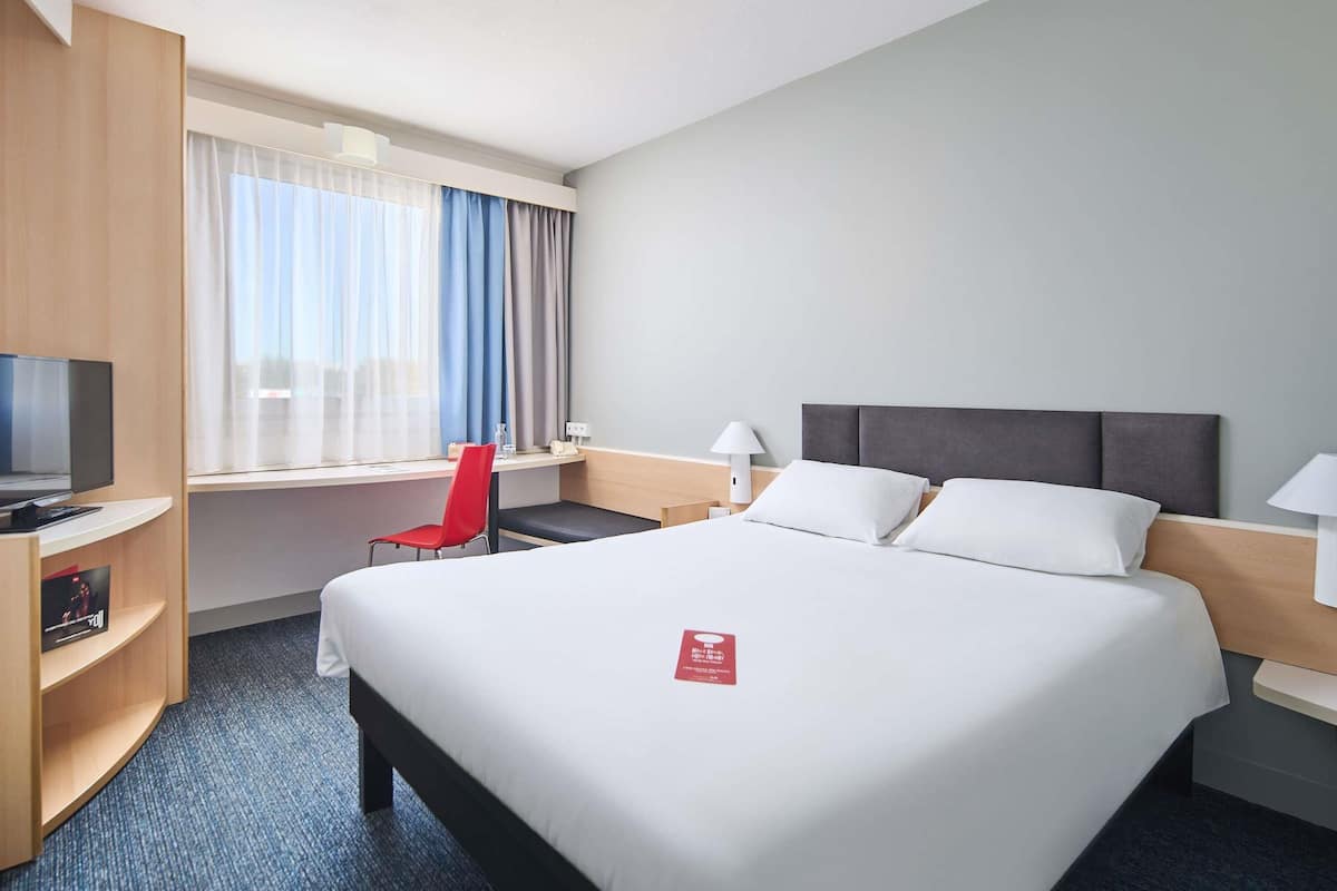 A hotel room with a large bed, a desk with a chair, a television, and a window with curtains.