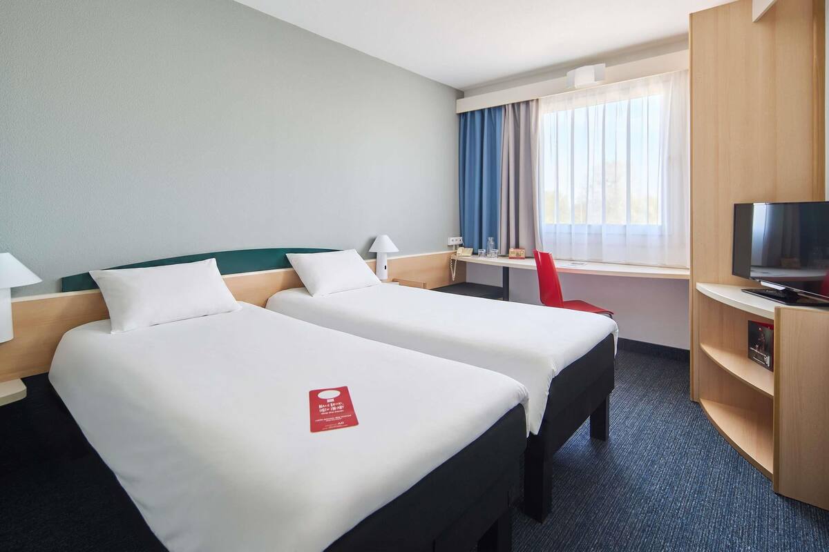 A hotel room with two beds, a desk, a TV, and a window with curtains.