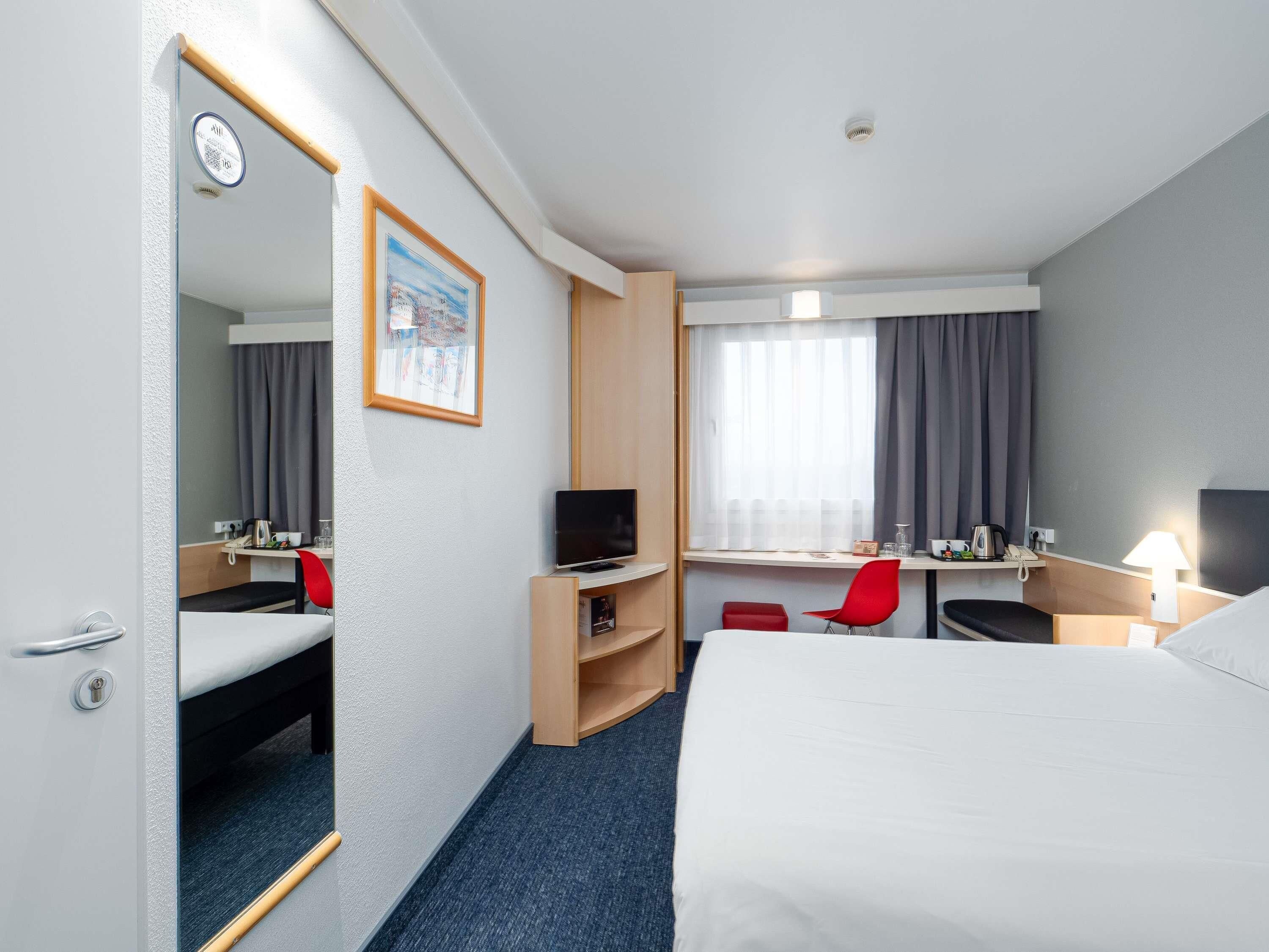 A hotel room with a bed, a desk with a computer, a chair, a TV, and a window with curtains.