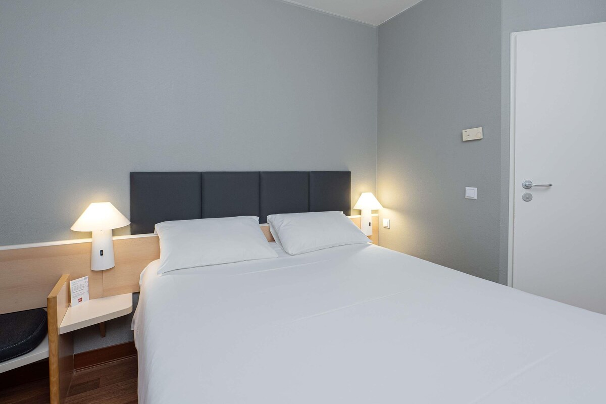 Una habitación de hotel con cama, mesitas de noche, lámparas y una puerta.