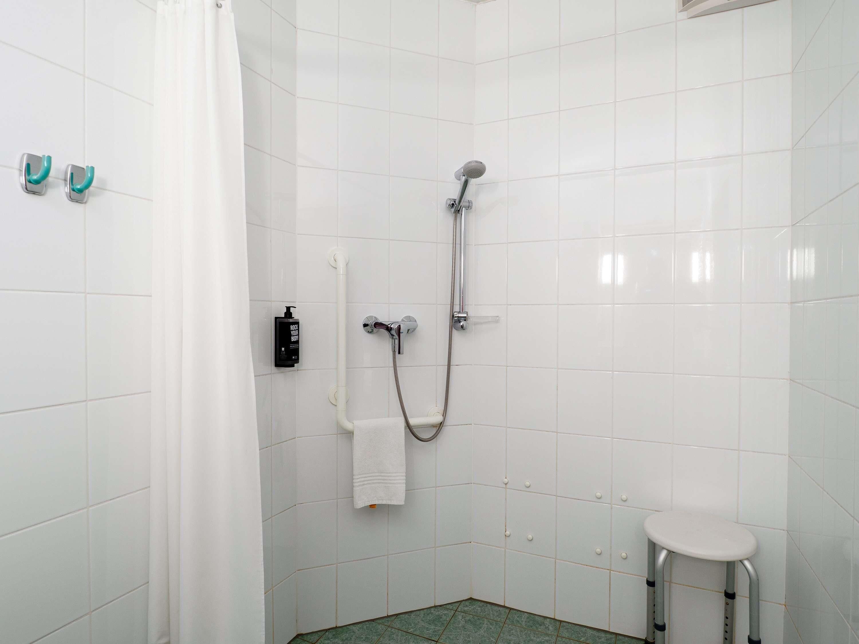 A small, white-tiled bathroom with a shower, a towel rack, a bottle of shampoo, and a small stool.