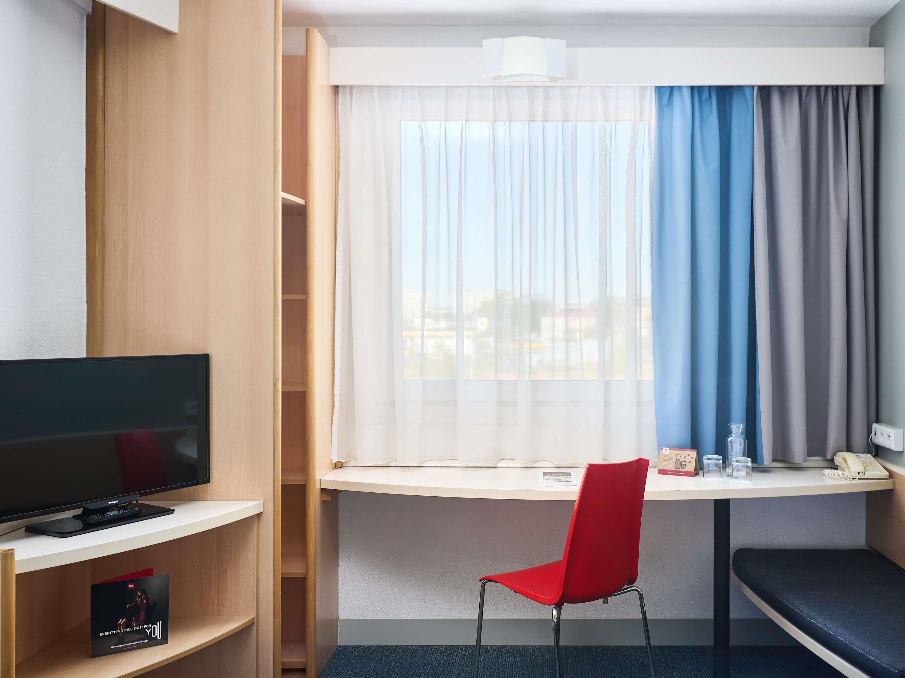 A hotel room with a TV, a desk, a red chair, a bench, and a window with curtains.