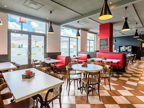 Un interior de restaurante con mesas, sillas y cabinas, ventanas amplias y lámparas colgantes.