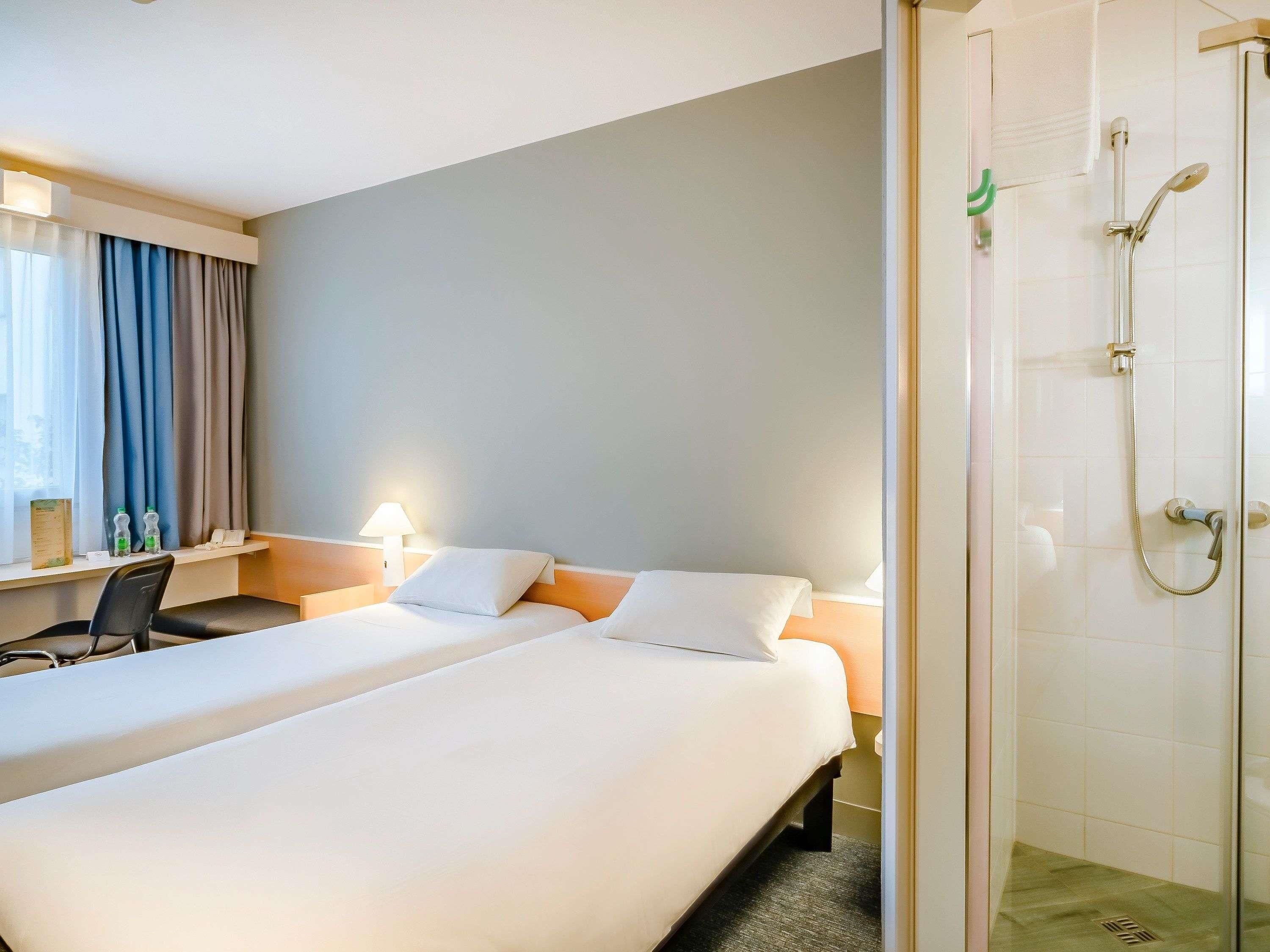 A hotel room with two beds, a desk, a chair, a lamp, and a shower area.