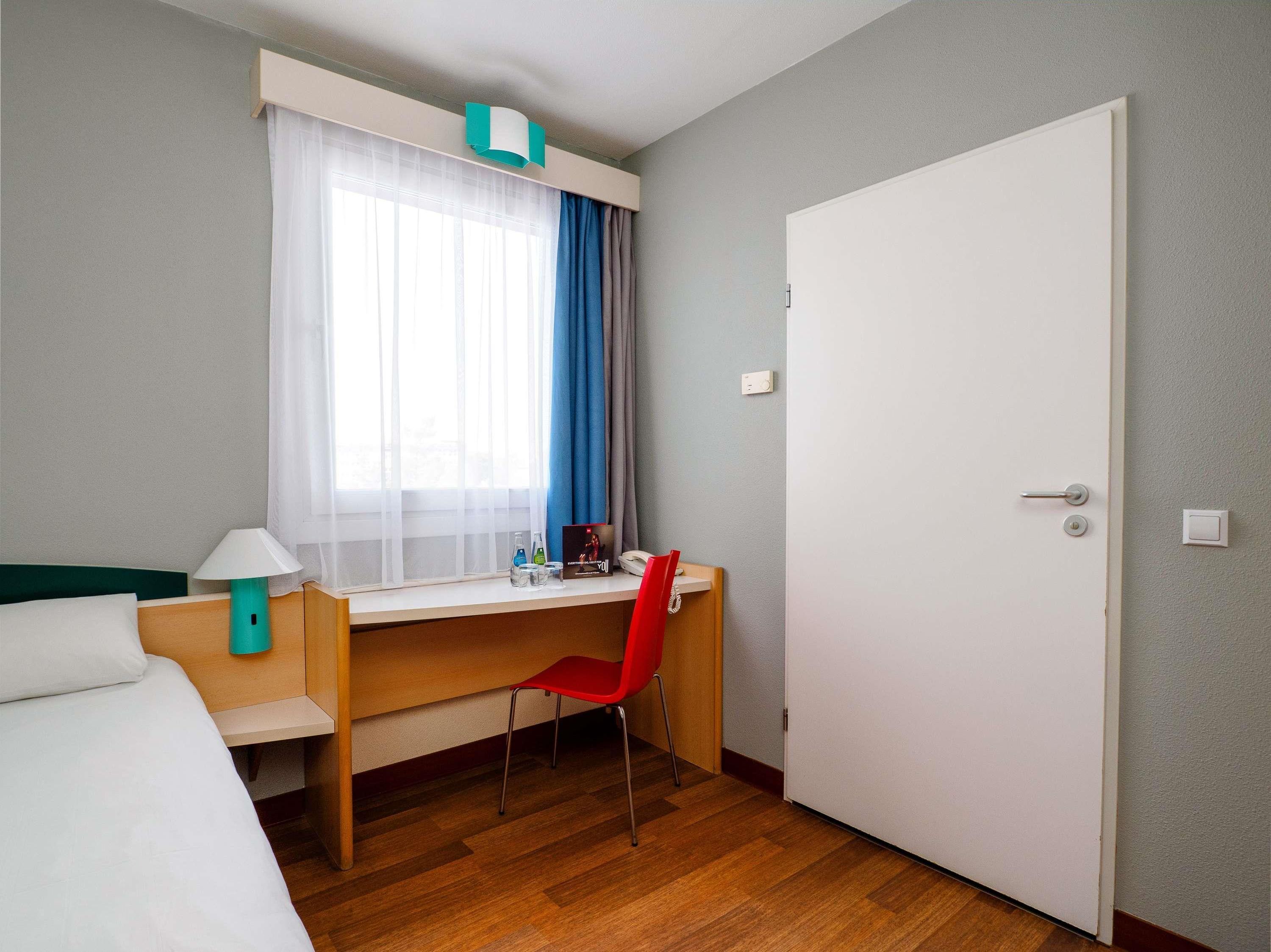 A hotel room with a bed, a desk with a chair, a window with curtains, and a door.