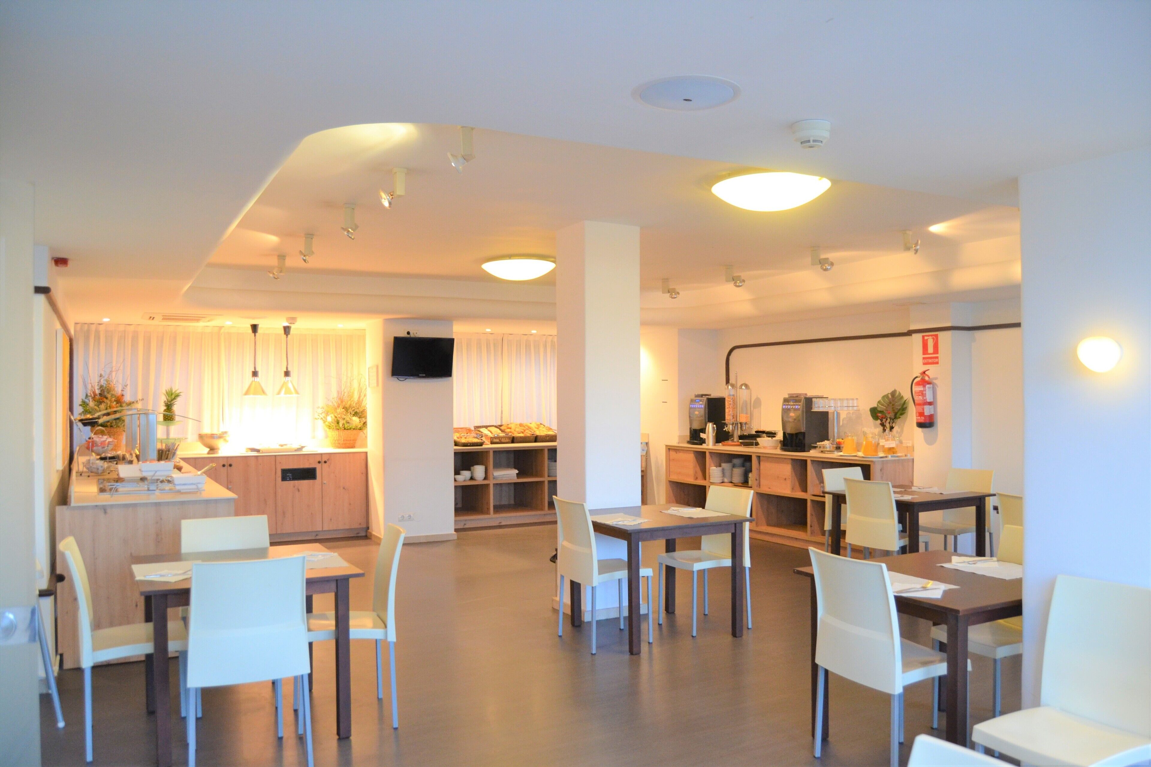 Een modern restaurantgedeelte in een hotel, met meerdere tafels en stoelen, een keuken met een koffieapparaat, en een televisie aan de muur.