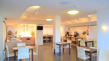 Een modern restaurantgedeelte in een hotel, met meerdere tafels en stoelen, een keuken met een koffieapparaat, en een televisie aan de muur.